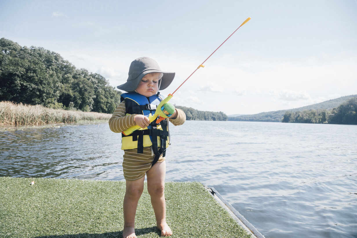 Cute baby boy with fishing rod standing by river on sunny day