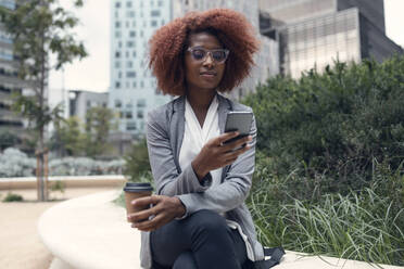 Young businesswoman sitting outdoors with smart phone and disposable coffee cup in hands - JSRF01946