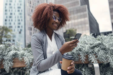 Young businesswoman sitting outdoors with smart phone and disposable coffee cup in hands - JSRF01947