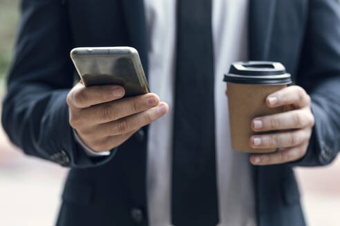 Hände eines Geschäftsmannes, der ein Smartphone benutzt und einen Einweg-Kaffeebecher hält - JSRF01964