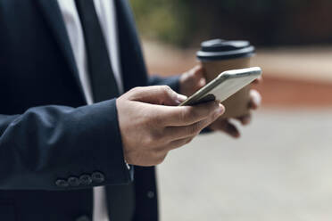Hände eines Geschäftsmannes, der ein Smartphone benutzt und einen Einweg-Kaffeebecher hält - JSRF01965
