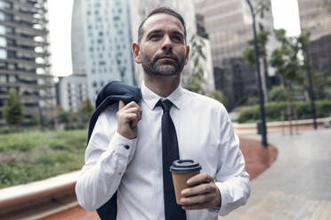 Portrait of businessman standing outdoors with jacket and disposable coffee cup in hands - JSRF01971