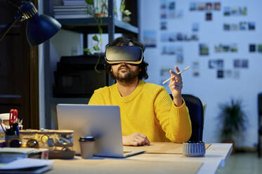 Businessman with laptop wearing virtual reality simulator sitting on chair in office - KIJF04406