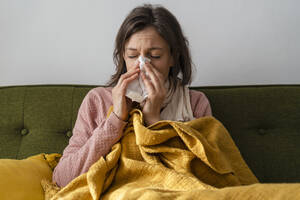 Sick woman blowing nose in facial tissue sitting on sofa at home - DIGF17732