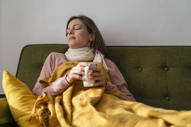 Sick woman holding tea cup resting on sofa at home - DIGF17734