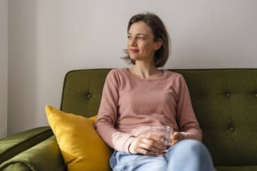 Smiling woman with glass of water sitting on sofa at home - DIGF17735