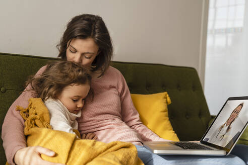 Mother embracing sick daughter consulting doctor online through laptop at home - DIGF17741