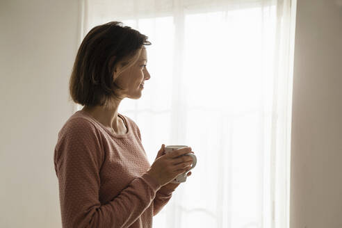 Lächelnde Frau mit Kaffeetasse, die zu Hause am Fenster steht - DIGF17743