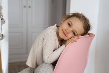Lächelndes süßes Mädchen sitzt auf rosa Stuhl zu Hause - DIGF17746