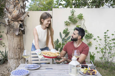 Happy woman showing homemade savory pie to man sitting at table in garden - SVKF00019