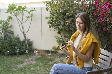 Lächelnde Frau mit langen Haaren, die im Garten sitzend ein Smartphone benutzt - SVKF00034