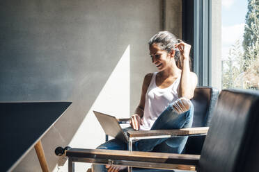 Smiling woman using laptop sitting on chair at home - JOSEF07973