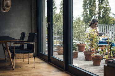 Woman using tablet PC sitting in balcony seen through glass window - JOSEF07978