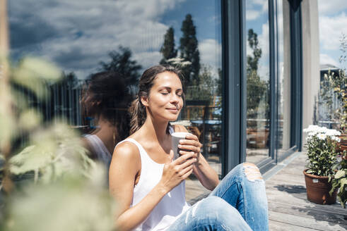 Smiling woman with eyes closed holding disposable coffee cup sitting in front of glass window at balcony - JOSEF07980