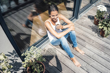 Happy woman with coffee cup sitting in balcony on sunny day - JOSEF07983