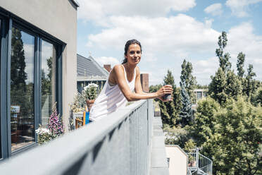 Lächelnde Frau, die eine Einweg-Kaffeetasse hält und sich an ein Balkongeländer lehnt, an einem sonnigen Tag - JOSEF07987