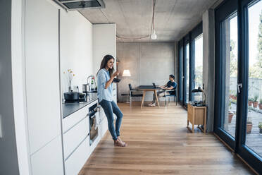 Woman with bowl standing in kitchen with man using laptop working from home - JOSEF07988