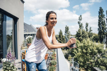 Happy beautiful woman with disposable coffee cup standing in balcony on sunny day - JOSEF07989