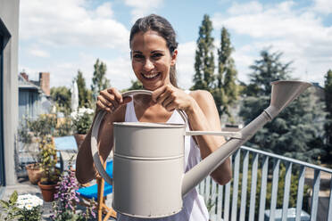 Cheerful woman holding watering can in balcony on sunny day - JOSEF07990