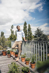 Young man standing on one leg leaning on railing on balcony on sunny day - JOSEF08003
