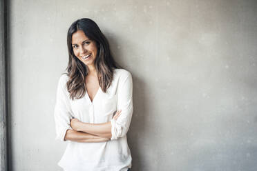 Cheerful woman standing with arms crossed in front of wall - JOSEF08018