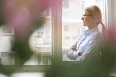 Businesswoman with arms crossed looking through window at work place - JOSEF08071