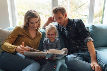 Parents reading book to daughter sitting on sofa in living room - JOSEF08089