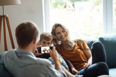 Happy blond woman and son photographed by man through smart phone in living room - JOSEF08092
