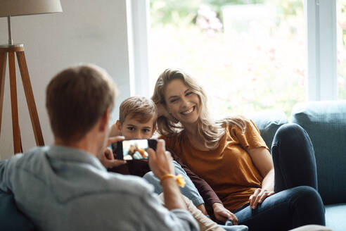 Glückliche blonde Frau und Sohn von Mann durch Smartphone im Wohnzimmer fotografiert - JOSEF08092