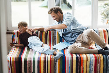 Glücklicher Vater, der seinen verspielten Sohn betrachtet, der ein Smartphone benutzt und zu Hause auf dem Sofa sitzt - JOSEF08099