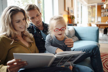 Father and mother looking at cute daughter reading book in living room at home - JOSEF08109