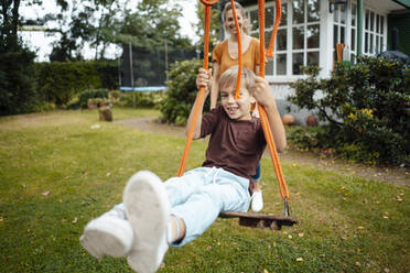 Smiling boy swinging in front of mother at backyard - JOSEF08116