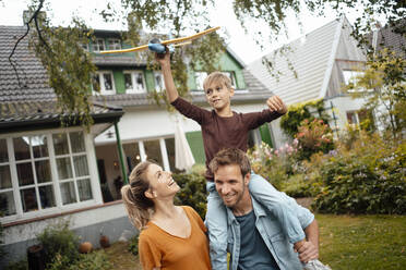 Happy woman looking at son holding airplane sitting on man shoulders at backyard - JOSEF08123