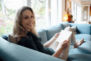 Happy beautiful blond woman sitting with tablet PC on sofa at home - JOSEF08124