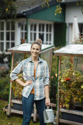 Happy blond woman with hand on hip holding watering can at backyard - JOSEF08129