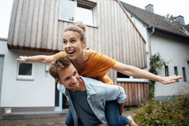 Happy man giving piggyback ride to woman with arms outstretched enjoying at backyard - JOSEF08140