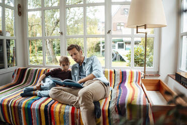 Vater liest seinem Sohn auf dem Sofa im Wohnzimmer zu Hause ein Buch vor - JOSEF08144