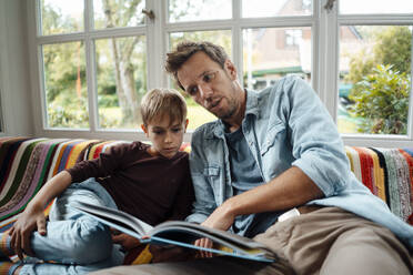 Man reading book to son sitting on sofa at home - JOSEF08145