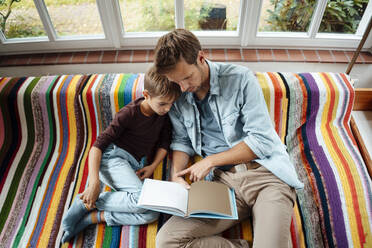 Vater unterrichtet Sohn beim Lesen eines Buches auf dem Sofa zu Hause - JOSEF08146