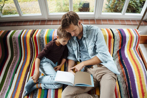 Vater unterrichtet Sohn beim Lesen eines Buches auf dem Sofa zu Hause - JOSEF08146