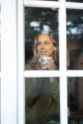 Beautiful blond woman holding coffee mug seen through glass window - JOSEF08148