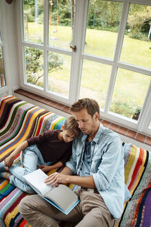 Mann unterrichtet Sohn beim Lesen eines Buches auf dem Sofa zu Hause - JOSEF08149