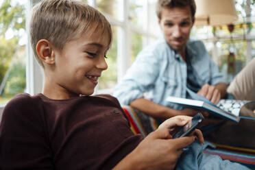 Glücklicher blonder Junge, der ein Smartphone benutzt und bei seinem Vater zu Hause sitzt - JOSEF08151