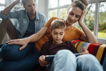 Mann und Frau sehen ihren Sohn an, der einen Tablet-PC benutzt und zu Hause auf dem Sofa sitzt - JOSEF08158