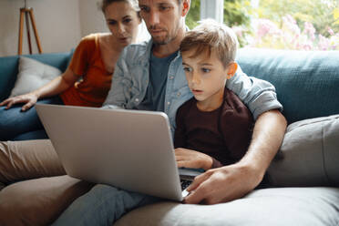 Vater und Sohn teilen sich einen Laptop und sitzen neben einer Frau auf dem Sofa zu Hause - JOSEF08166