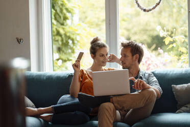 Woman holding credit card sitting by boyfriend with laptop on sofa in living room - JOSEF08169
