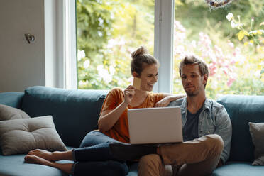 Cheerful woman holding credit card sitting by boyfriend using laptop on sofa in living room - JOSEF08170