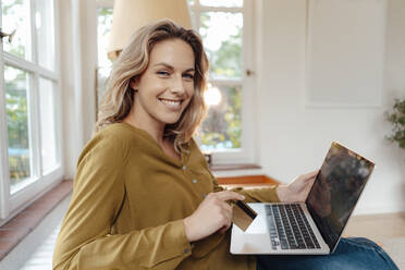 Glückliche blonde Frau mit Kreditkarte und Laptop zu Hause sitzend - JOSEF08188