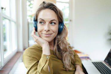 Smiling beautiful woman listening music through wireless headphones at home - JOSEF08189