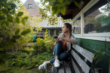 Woman with smart phone sitting on bench at backyard - JOSEF08197